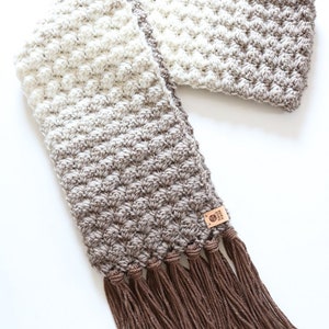 Puede incluir: Una bufanda de ganchillo con un patr&oacute;n de degradado marr&oacute;n y blanco y borlas marrones. La bufanda tiene una peque&ntilde;a etiqueta de cuero con el texto "Crochet by Amy".