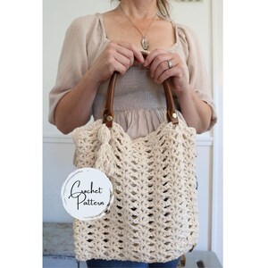 May include: A cream-colored crochet tote bag featuring an openwork stitch pattern, two brown leather handles attached with gold-tone metal rivets, and a matching crochet tassel. A circular white label with the text Crochet Pattern is attached.