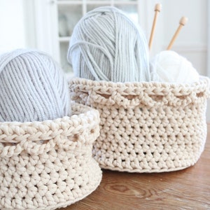 Puede incluir: Dos cestas de ganchillo color crema sobre una superficie de madera. Una cesta contiene una bola de hilo gris. La cesta m&aacute;s grande contiene una bola de hilo gris, hilo blanco y agujas de tejer de madera. Las cestas tienen un borde festoneado decorativo.