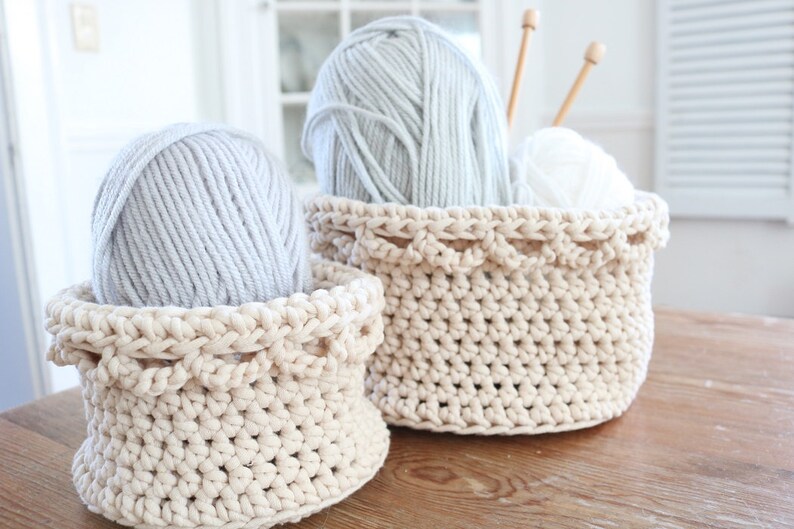 Puede incluir: Dos cestas de ganchillo color crema sobre una superficie de madera. Una cesta contiene una bola de hilo gris. La cesta m&aacute;s grande contiene una bola de hilo gris, hilo blanco y agujas de tejer de madera. Las cestas tienen un borde festoneado decorativo.