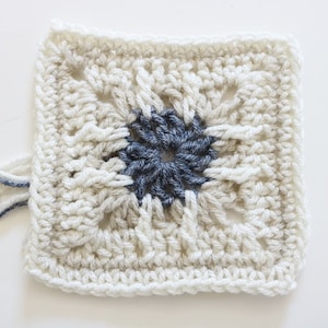 May include: A crocheted square with a white background and a dark blue center. The square has a textured pattern and is made with yarn.