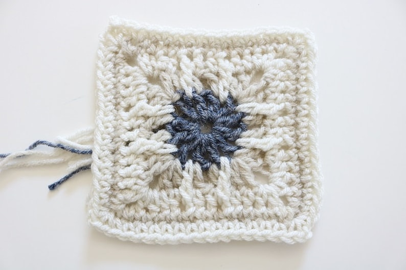 May include: A crocheted square with a white background and a dark blue center. The square has a textured pattern and is made with yarn.