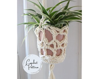 Puede incluir: Un colgador de plantas con un diseño de macramé color crema. El colgador sostiene una maceta de cerámica rosa llena de una planta verde y blanca. La parte inferior del macramé tiene una borla. La imagen incluye el texto "Crochet Pattern".