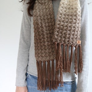 Puede incluir: Una bufanda de crochet color marr&oacute;n y crema con flecos marrones. La bufanda la lleva puesta una persona que lleva una camisa de manga larga gris y unos vaqueros azules.