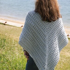 May include: A light blue crochet poncho with a lacy pattern. The poncho is worn by a person standing in a grassy field with a body of water in the background.