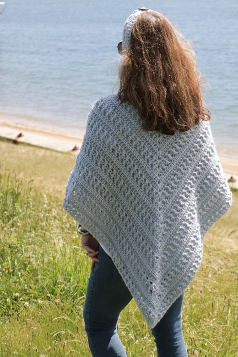 May include: A light blue crochet poncho with a lacy pattern. The poncho is worn by a person standing in a grassy field with a body of water in the background.