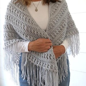 May include: A gray crocheted shawl with fringe. The shawl is draped over a person's shoulders and is being held in place by their hands.