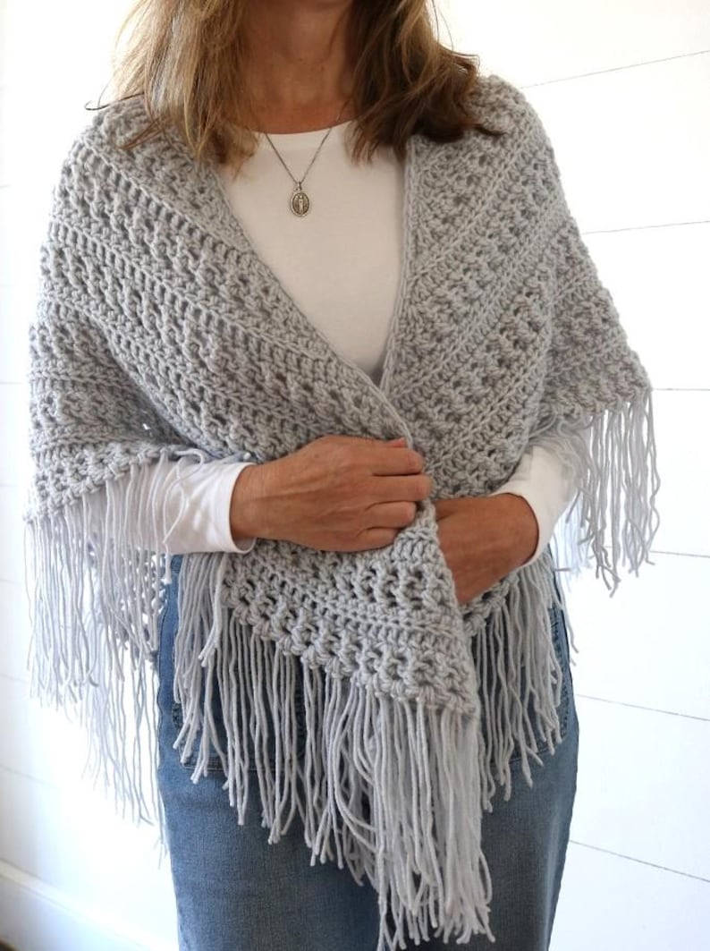 May include: A gray crocheted shawl with fringe. The shawl is draped over a person's shoulders and is being held in place by their hands.