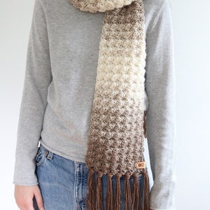 Puede incluir: Una bufanda de crochet color marr&oacute;n y crema con flecos marrones. La bufanda la lleva puesta una persona que lleva un jersey gris y unos vaqueros azules.