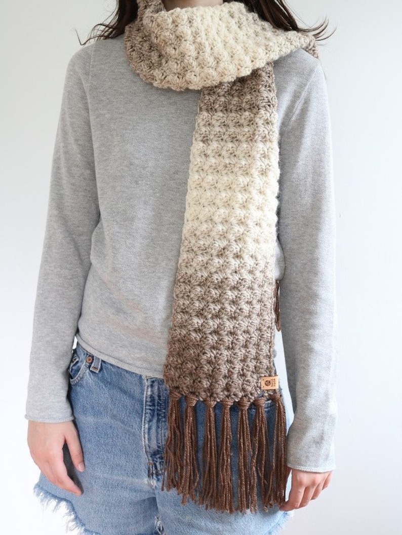 Puede incluir: Una bufanda de crochet color marr&oacute;n y crema con flecos marrones. La bufanda la lleva puesta una persona que lleva un jersey gris y unos vaqueros azules.