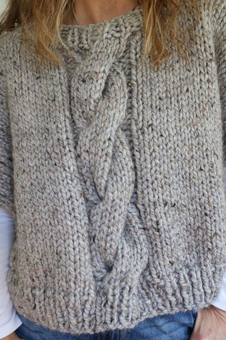Puede incluir: Un grueso su&eacute;ter de punto en color gris claro con un dise&ntilde;o de punto trenzado en la parte delantera. El su&eacute;ter tiene cuello redondo y mangas largas. El hilo tiene motas marrones y beige.