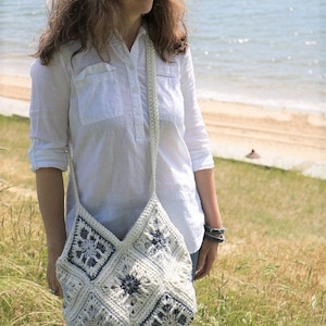 May include: A woman wearing a white button-down shirt and blue jeans is carrying a white crocheted tote bag with blue geometric designs. The bag is worn over the shoulder and has a long strap.