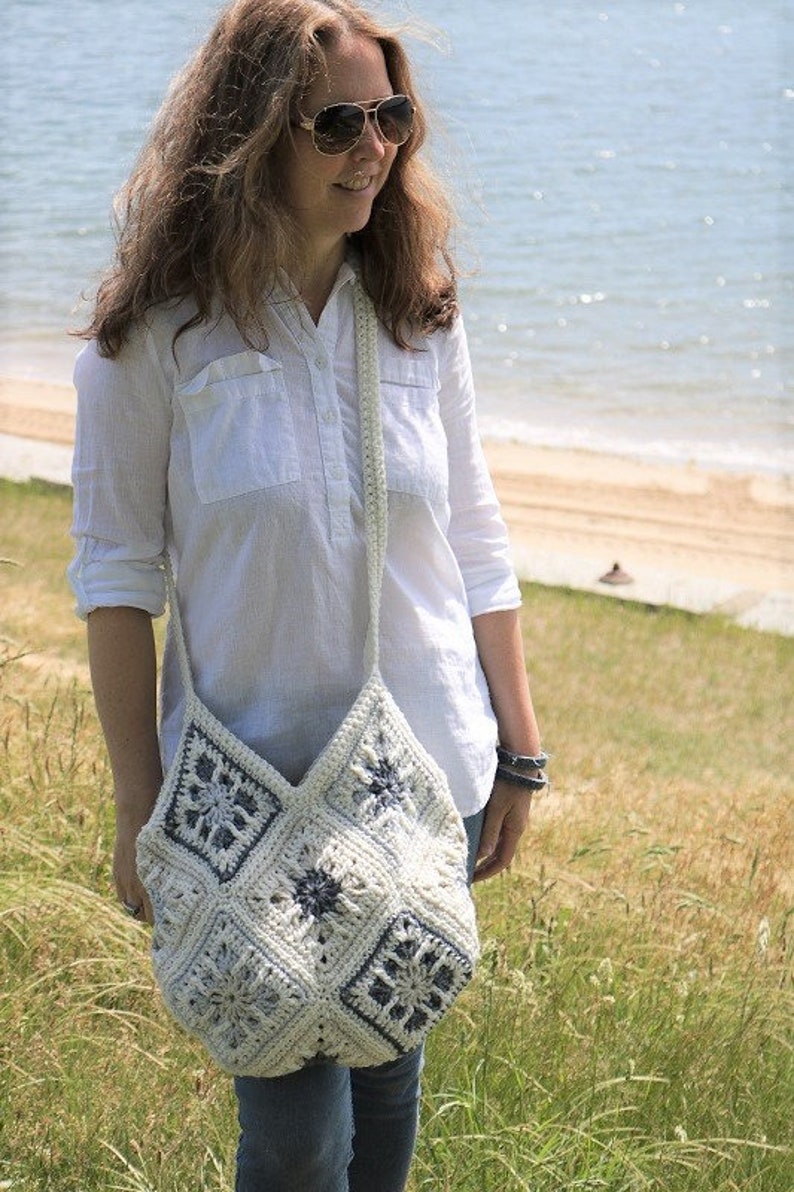 May include: A woman wearing a white button-down shirt and blue jeans is carrying a white crocheted tote bag with blue geometric designs. The bag is worn over the shoulder and has a long strap.