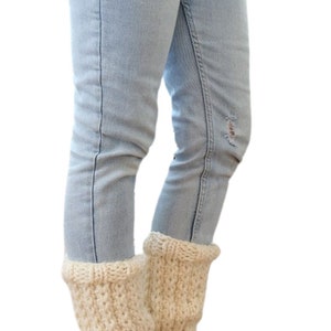 May include: A pair of cream-colored knitted leg warmers with fringe on the bottom. The leg warmers are worn over blue jeans and brown boots.