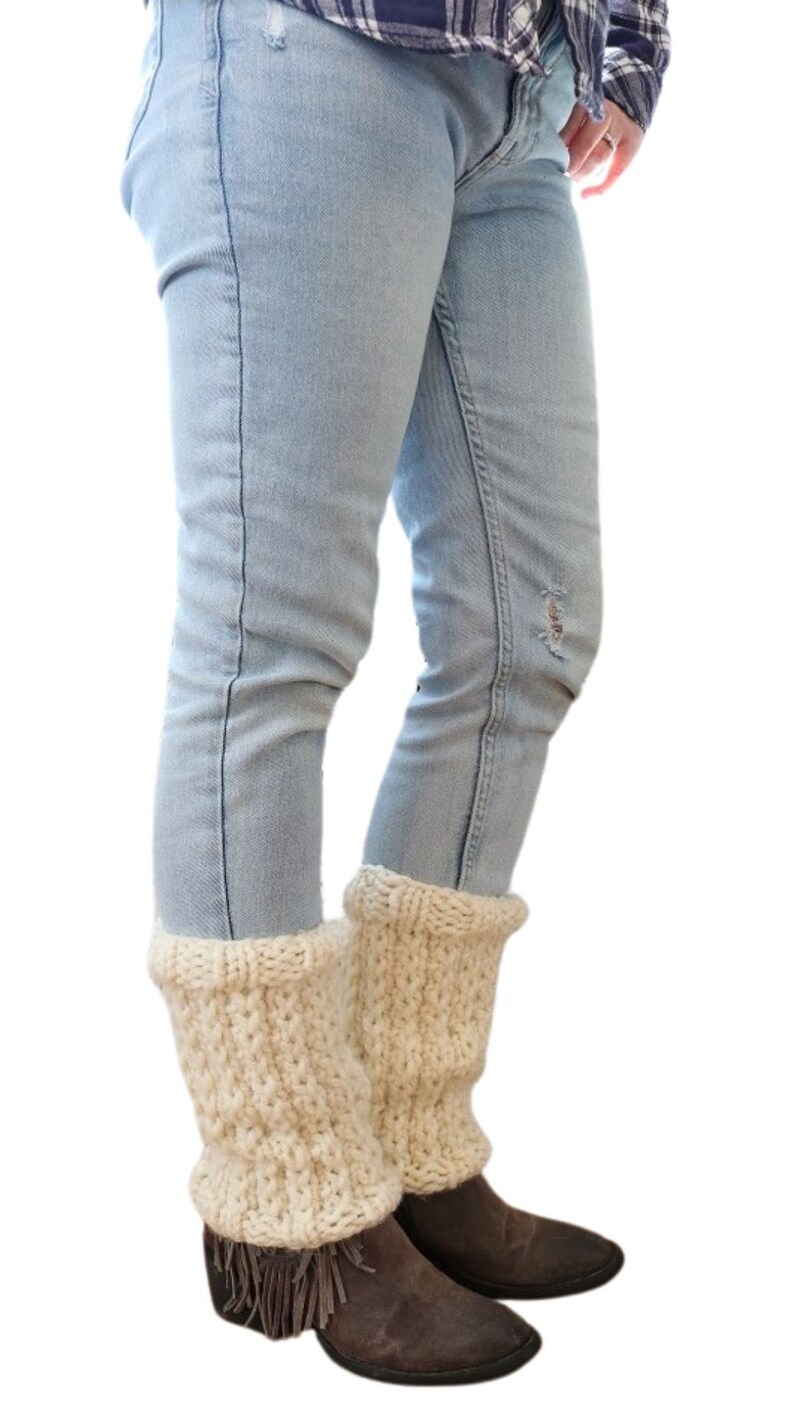 May include: A pair of cream-colored knitted leg warmers with fringe on the bottom. The leg warmers are worn over blue jeans and brown boots.
