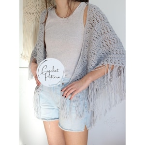 Può includere: Un scialle di uncinetto grigio chiaro con frange. Lo scialle è indossato sopra un top grigio senza maniche e pantaloncini di jeans. Il testo "Crochet Pattern" è scritto in un cerchio sull'immagine.