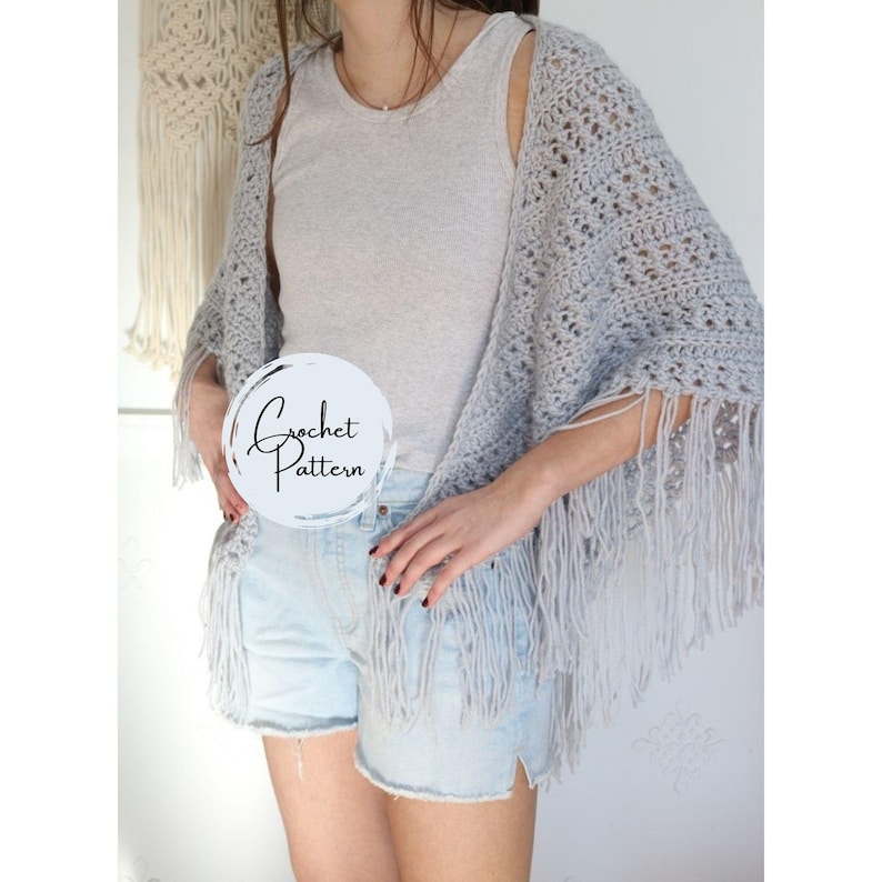 May include: A light gray crochet shawl with fringe. The shawl is worn over a gray tank top and denim shorts. The text "Crochet Pattern" is written in a circle on the image.