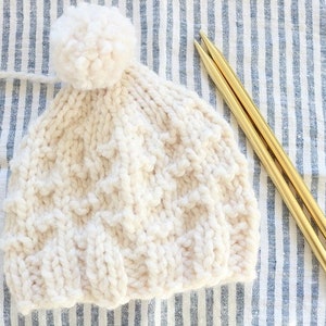 May include: A white knitted hat with a white pom pom on top. The hat is lying on a blue and white striped fabric with two gold knitting needles.