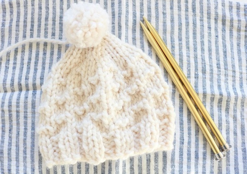 May include: A white knitted hat with a white pom pom on top. The hat is lying on a blue and white striped fabric with two gold knitting needles.