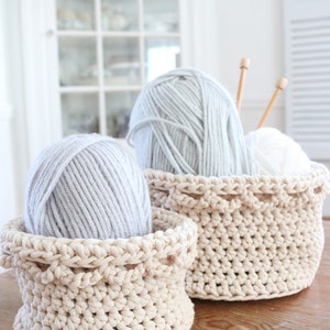Puede incluir: Dos cestas de ganchillo beige llenas de hilo gris y blanco. Agujas de tejer de madera est&aacute;n en una cesta. Las cestas est&aacute;n sobre una superficie de madera, con un armario blanco al fondo. El hilo es suave y esponjoso.