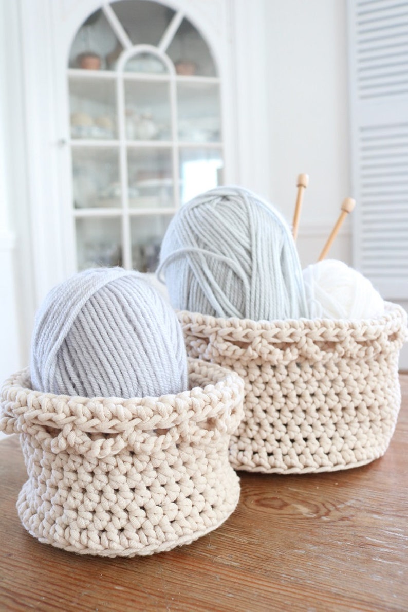 Puede incluir: Dos cestas de ganchillo beige llenas de hilo gris y blanco. Agujas de tejer de madera est&aacute;n en una cesta. Las cestas est&aacute;n sobre una superficie de madera, con un armario blanco al fondo. El hilo es suave y esponjoso.