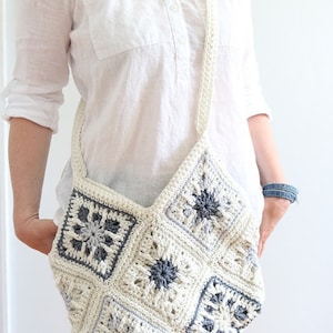 May include: A white crocheted bag with a gray and white geometric pattern. The bag has a long strap and is worn over the shoulder.