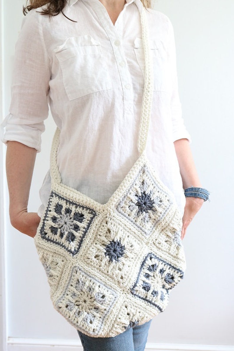 May include: A white crocheted bag with a gray and white geometric pattern. The bag has a long strap and is worn over the shoulder.