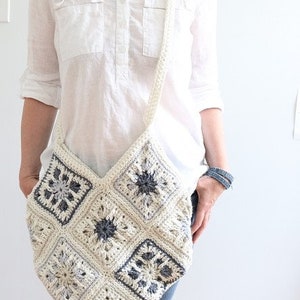 May include: A white crocheted shoulder bag with a geometric pattern in shades of gray. The bag is worn over the shoulder of a person wearing a white shirt and blue jeans.