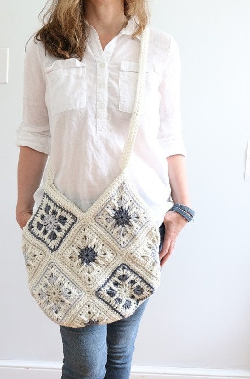 May include: A white crocheted shoulder bag with a geometric pattern in shades of gray. The bag is worn over the shoulder of a person wearing a white shirt and blue jeans.