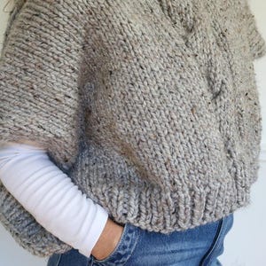 Puede incluir: Un poncho de punto grueso en color gris claro con motas de tonos m&aacute;s oscuros. El poncho tiene un cuello ancho y mangas cortas. La modelo lleva una camisa blanca de manga larga y vaqueros azules.