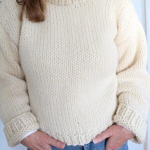 May include: Cream-colored, chunky knit sweater with a ribbed crew neck and cuffs. The sweater has long sleeves and a relaxed fit. The person is wearing blue jeans. The sweater is made of a thick, warm yarn, perfect for cooler weather.