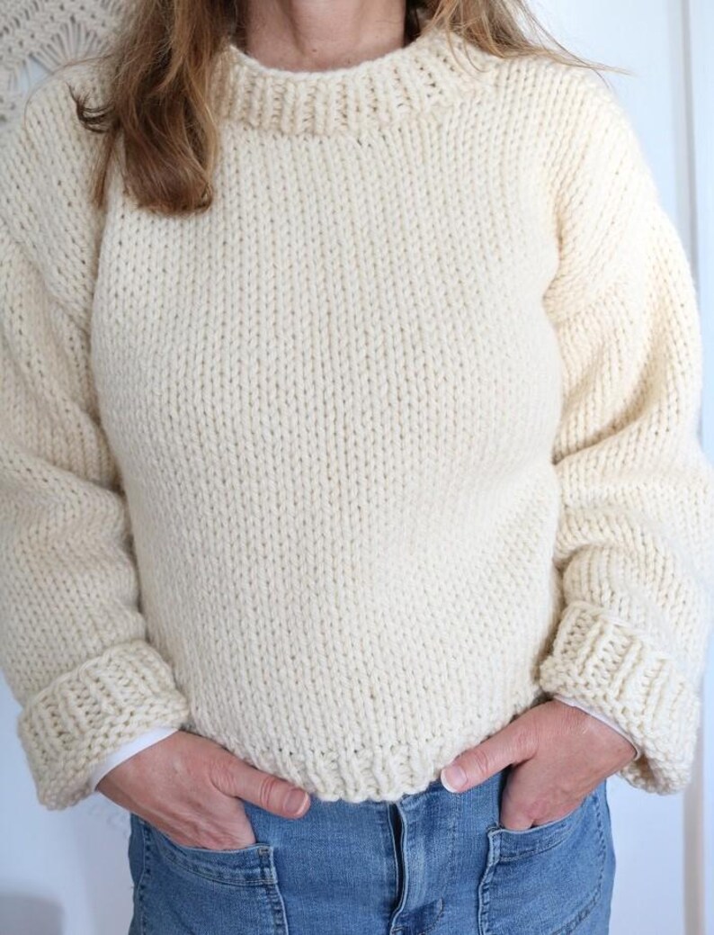 May include: Cream-colored, chunky knit sweater with a ribbed crew neck and cuffs. The sweater has long sleeves and a relaxed fit. The person is wearing blue jeans. The sweater is made of a thick, warm yarn, perfect for cooler weather.