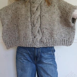 Puede incluir: Un poncho de punto grueso gris con un dise&ntilde;o de punto trenzado en la parte delantera. El poncho tiene un cuello redondo y mangas cortas. La tela tiene motas negras. La persona de la imagen lleva vaqueros azules.