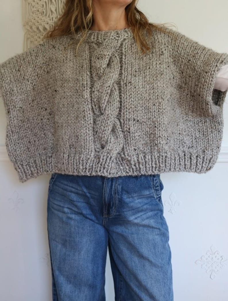 Puede incluir: Un poncho de punto grueso gris con un dise&ntilde;o de punto trenzado en la parte delantera. El poncho tiene un cuello redondo y mangas cortas. La tela tiene motas negras. La persona de la imagen lleva vaqueros azules.