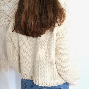 May include: Cream-colored, chunky knit sweater with a cropped hem. The sweater is paired with blue jeans. A tortoiseshell hair clip is visible in the model's brown hair. A macrame wall hanging is in the background.