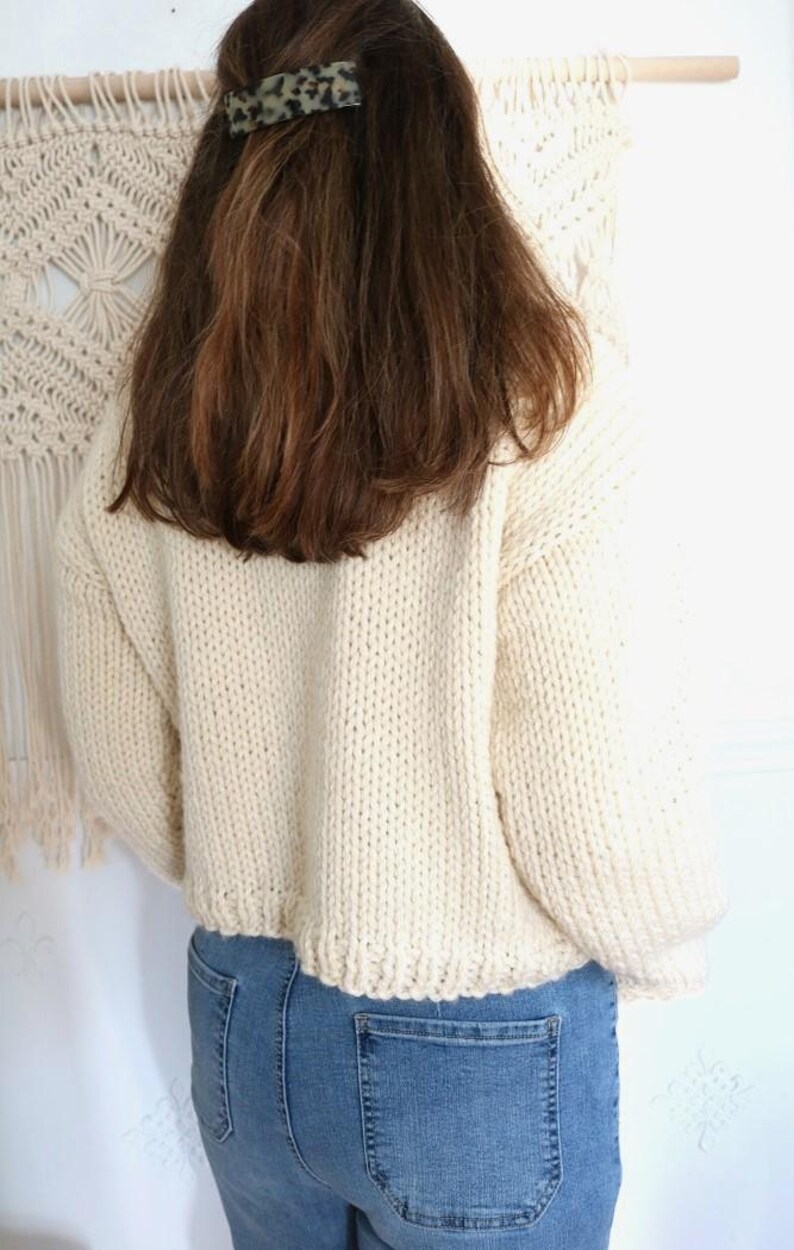 May include: Cream-colored, chunky knit sweater with a cropped hem. The sweater is paired with blue jeans. A tortoiseshell hair clip is visible in the model's brown hair. A macrame wall hanging is in the background.
