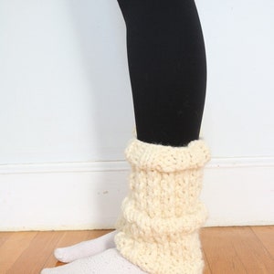 May include: Cream-colored knitted leg warmers with a ribbed texture. The leg warmers are worn over white socks and black leggings.