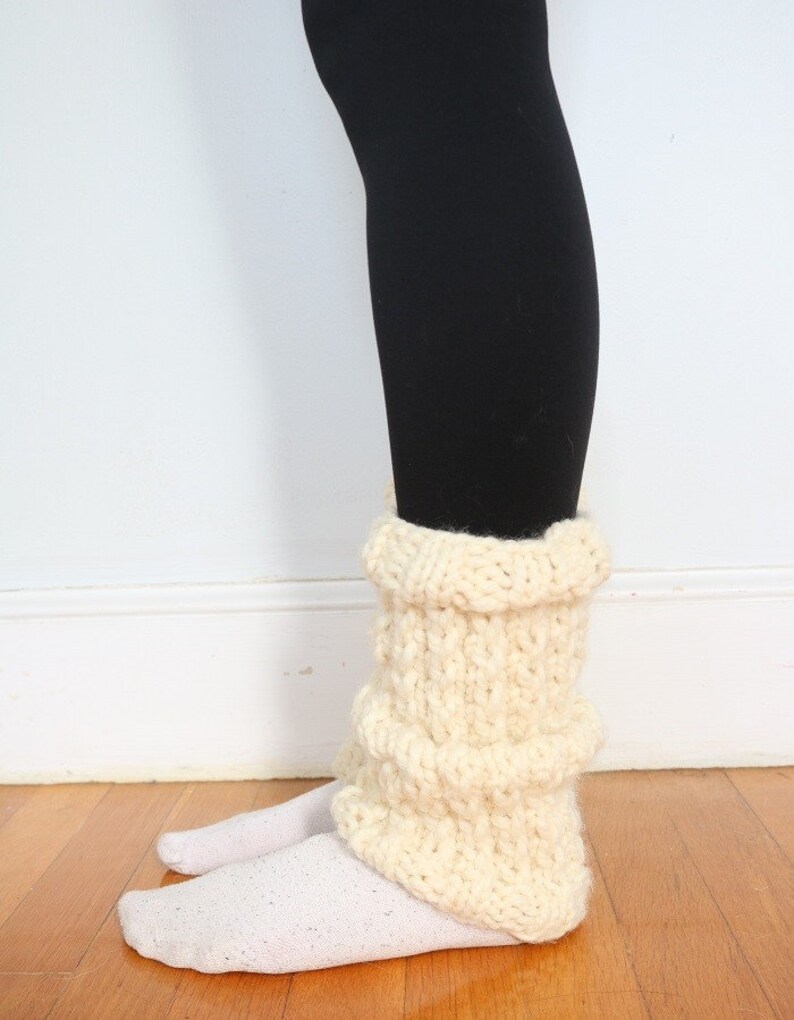 May include: Cream-colored knitted leg warmers with a ribbed texture. The leg warmers are worn over white socks and black leggings.