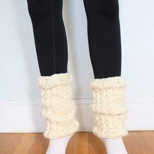 May include: A pair of white knitted leg warmers with a textured pattern. The leg warmers are worn over black leggings.