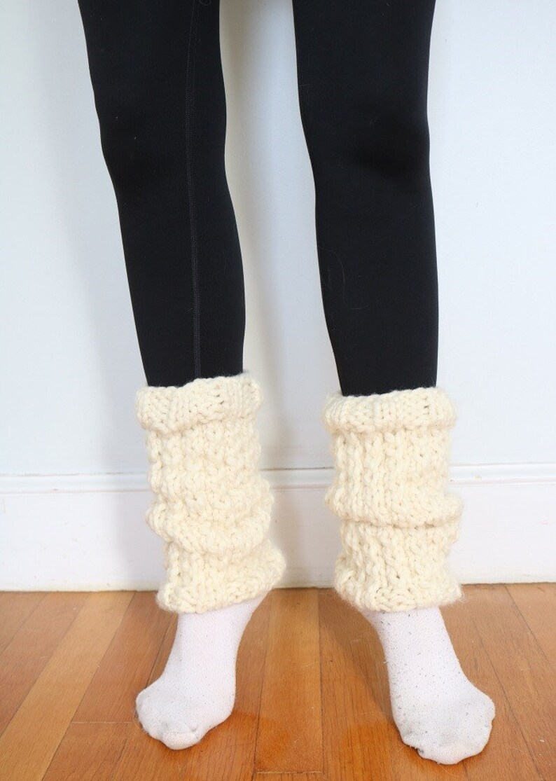 May include: A pair of white knitted leg warmers with a textured pattern. The leg warmers are worn over black leggings.