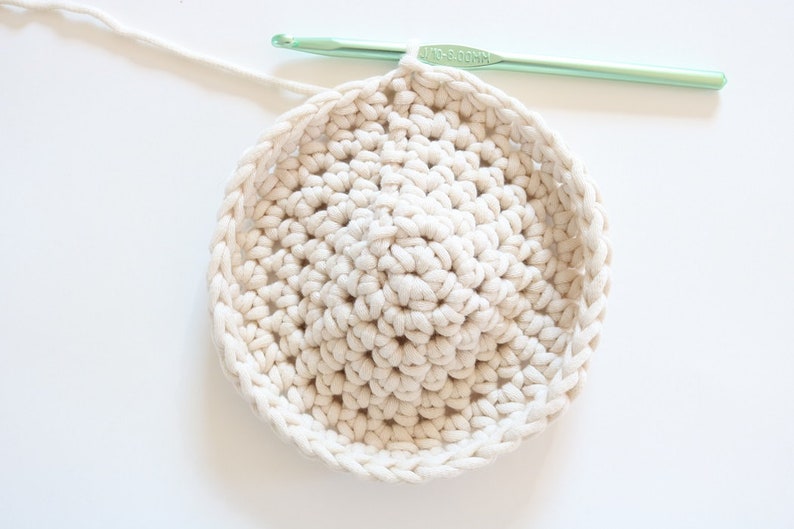 Puede incluir: Primer plano de un proyecto de ganchillo circular de color crema en curso. Un ganchillo verde claro est&aacute; posicionado cerca del borde del trabajo, con un hilo blanco que se extiende desde el proyecto.