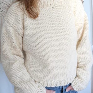 May include: Cream-colored, hand-knitted sweater with a crew neck and long sleeves. The sweater has a thick, textured knit and a ribbed hem and cuffs. The style is a classic, oversized fit. The sweater is made of a soft, warm yarn.