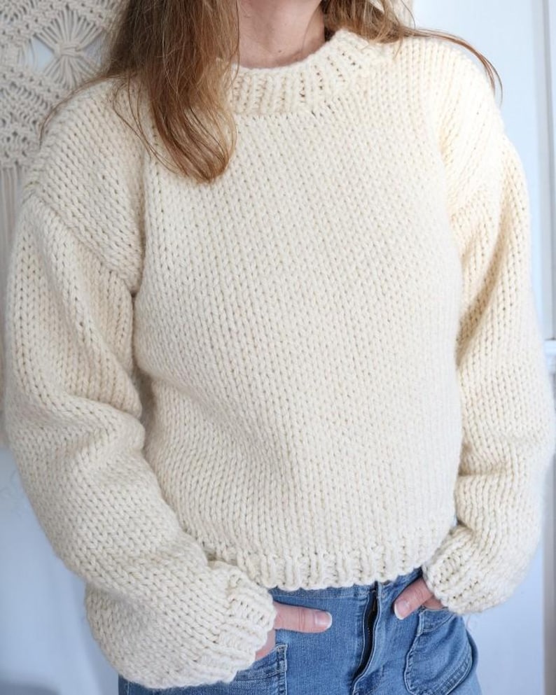 May include: Cream-colored, hand-knitted sweater with a crew neck and long sleeves. The sweater has a thick, textured knit and a ribbed hem and cuffs. The style is a classic, oversized fit. The sweater is made of a soft, warm yarn.