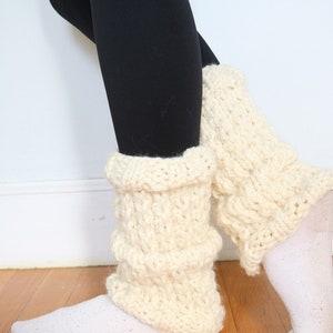 May include: A pair of white knitted leg warmers with a chunky knit texture. The leg warmers are worn over black leggings.