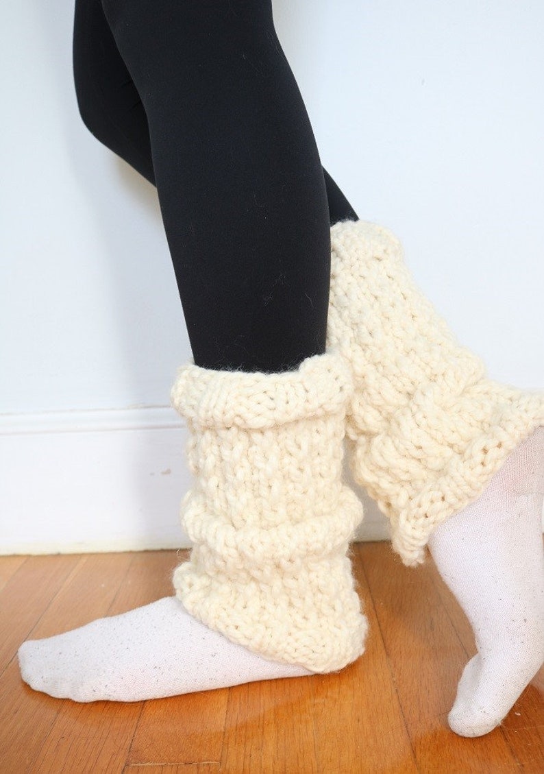 May include: A pair of white knitted leg warmers with a chunky knit texture. The leg warmers are worn over black leggings.