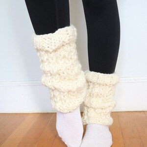 May include: A pair of white knitted leg warmers with a textured pattern. The leg warmers are worn over white socks and black leggings.