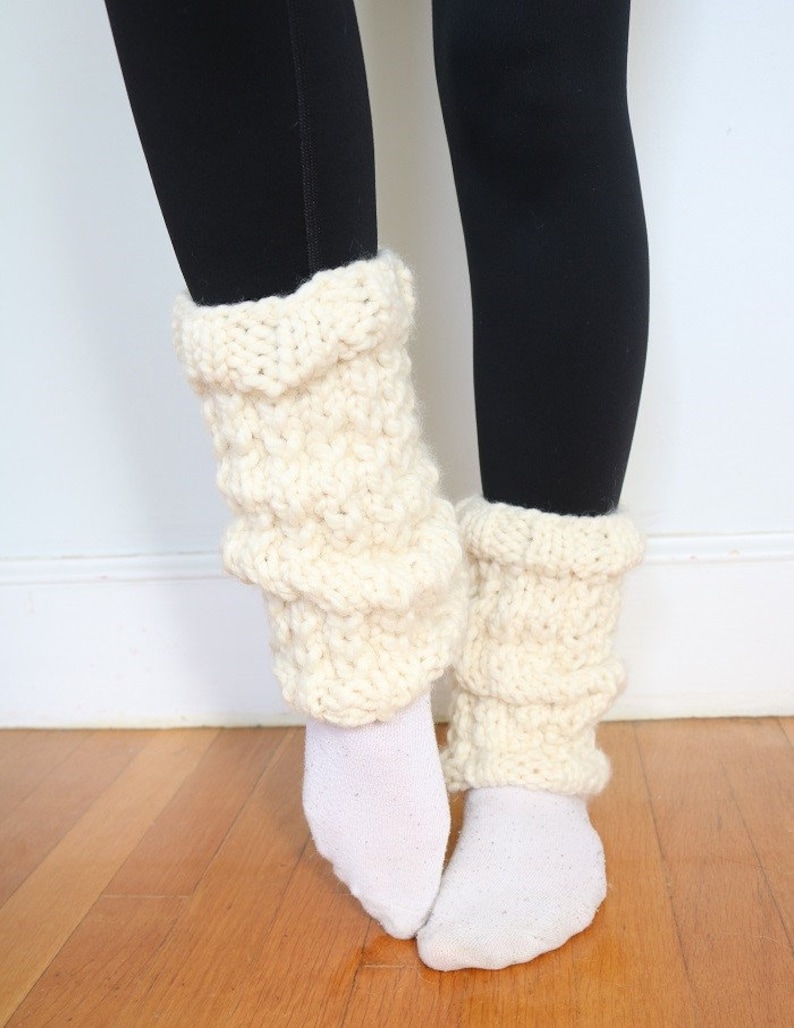 May include: A pair of white knitted leg warmers with a textured pattern. The leg warmers are worn over white socks and black leggings.