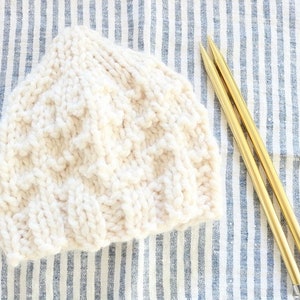May include: A white knitted hat with a textured pattern, lying on a blue and white striped fabric with two gold knitting needles.