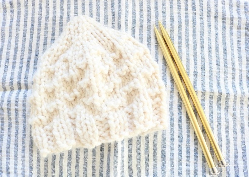 May include: A white knitted hat with a textured pattern, lying on a blue and white striped fabric with two gold knitting needles.