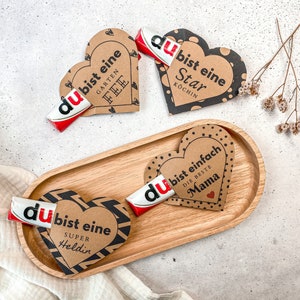 May include: Three brown paper heart-shaped gift tags with black polka dots and black text. Each tag has a chocolate bar tucked inside. The text on the tags reads: "du bist eine Garten Fee", "du bist eine Star Kochlin", and "du bist einfach die beste Mama".
